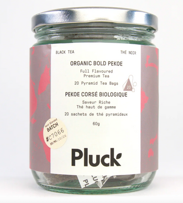A glass jar of Pluck Organic Bold Pekoe tea bags, with a silver lid and a pink and red patterned label. The label states